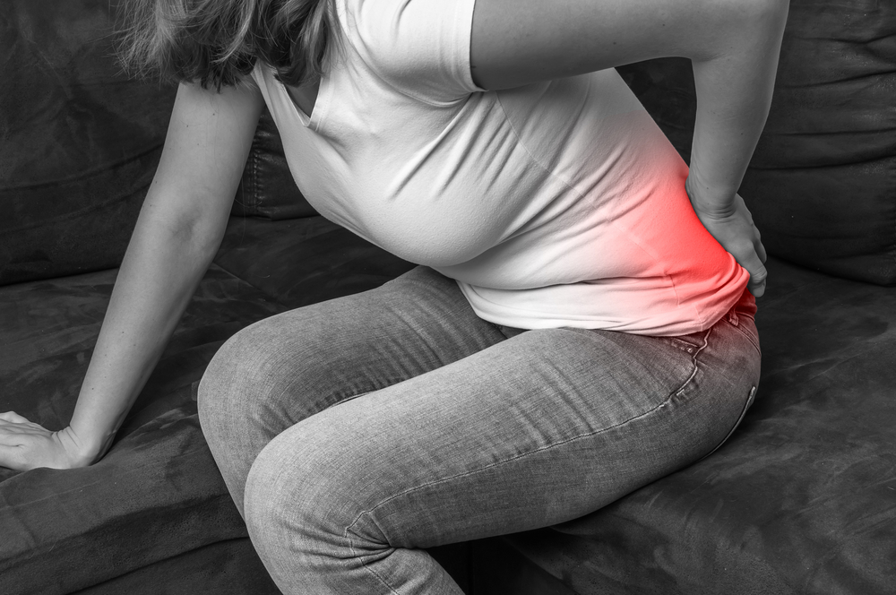 A woman sits on a couch holding her lower back in pain, highlighted by a red area. Wearing a white t-shirt and jeans, she illustrates the need for orthopedic care or Pelvic Floor Physical Therapy at Home. The image is mostly black and white except for the red.