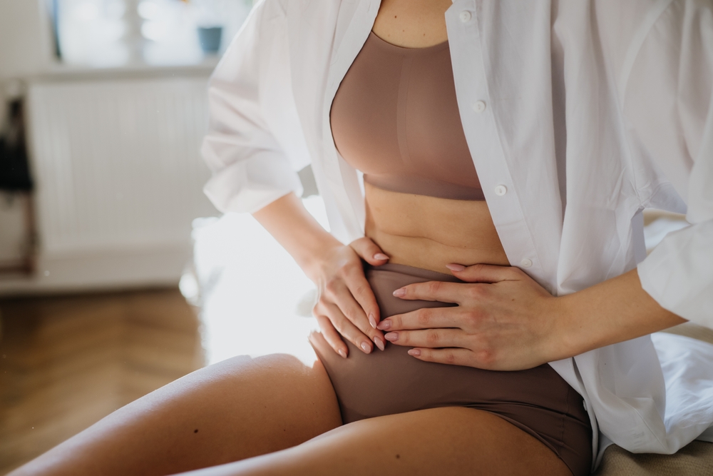 A person wearing a white shirt and beige underwear sits indoors, holding their hands on their lower abdomen, possibly indicating discomfort—an experience often related to pelvic health or pregnancy & postpartum concerns.