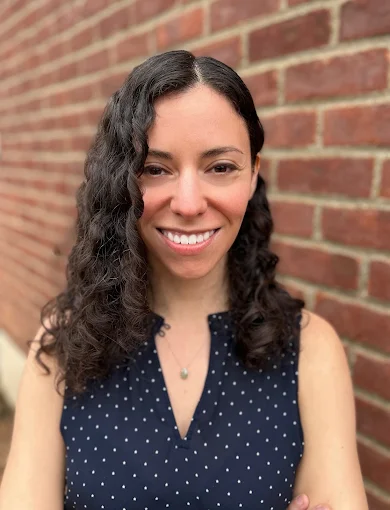 A woman with long, curly dark hair is smiling and standing in front of a red brick wall. She is wearing a navy blue sleeveless top with white polka dots, a small necklace, and radiates confidence in her journey toward pelvic health.