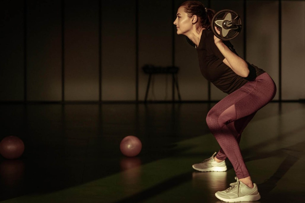 A woman wearing a black top and purple leggings performs a squat with a barbell on her shoulders in a dimly lit gym, highlighting the importance of pelvic health, with two pink exercise balls on the floor nearby.